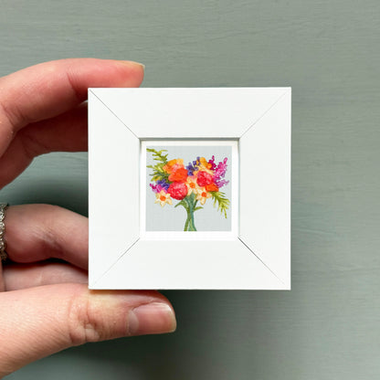 A hand is holding a small white square frame containing a colorful painting of flowers.