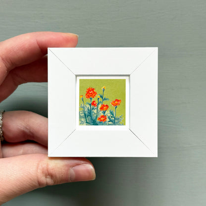 A hand is holding a small white square frame containing a painting of orange flowers against a green background.