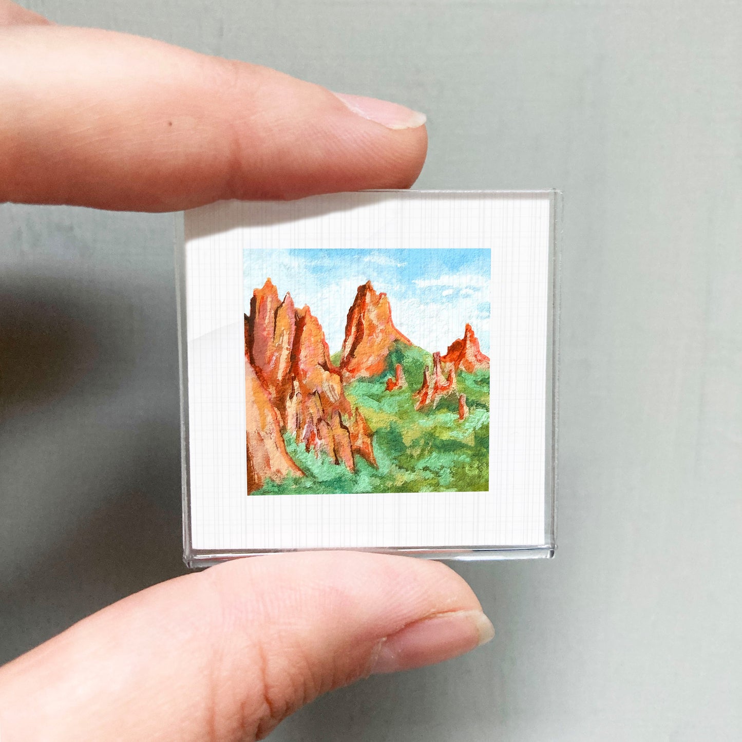 A person's hand holding a small square picture of a mountain landscape with red rock formations and green vegetation.