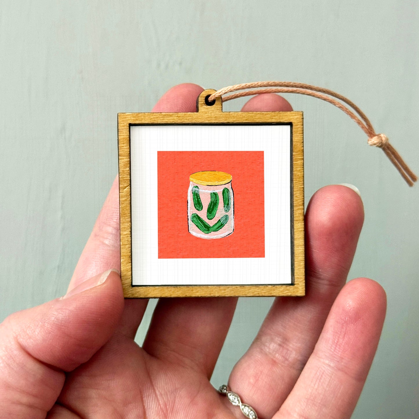 A hand holding a small wooden frame containing a picture of a jar of pickles.