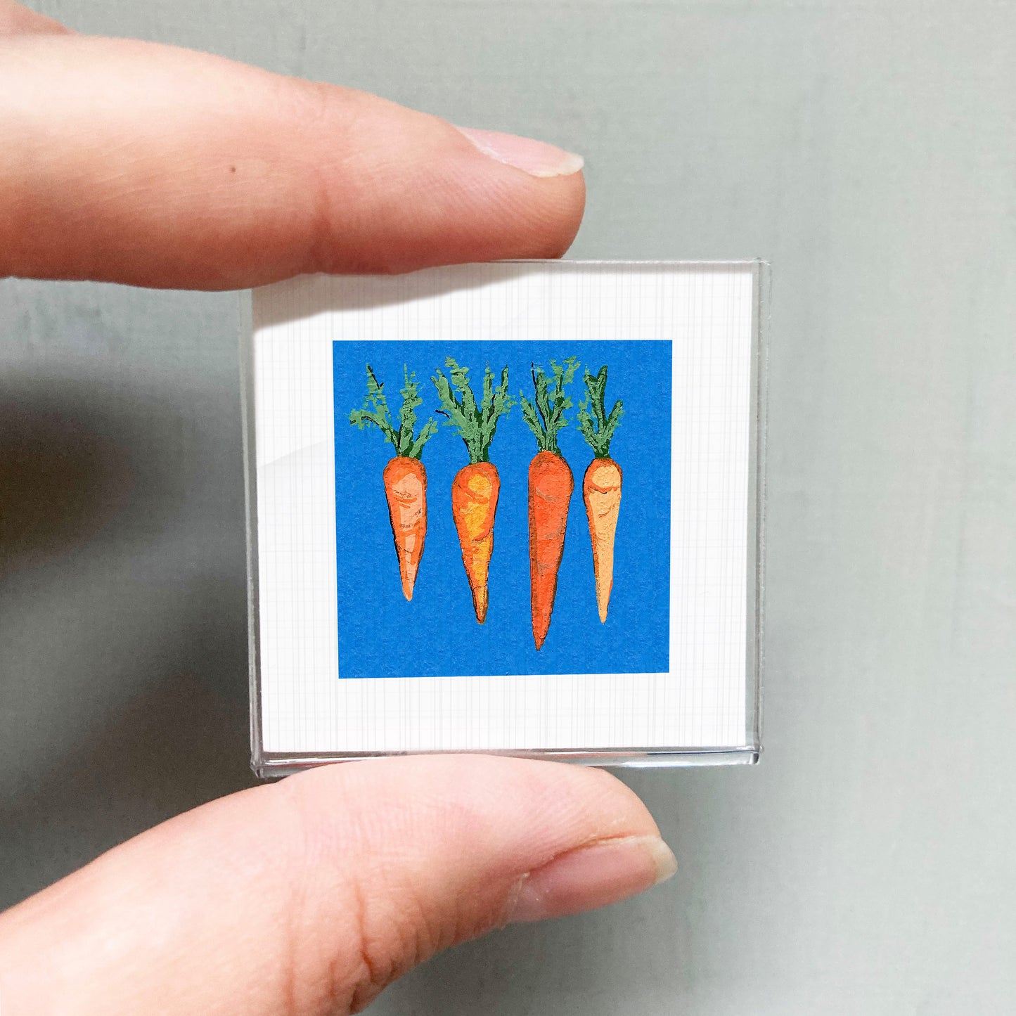 A person's hand is holding a small square frame containing an image of four carrots against a blue background.