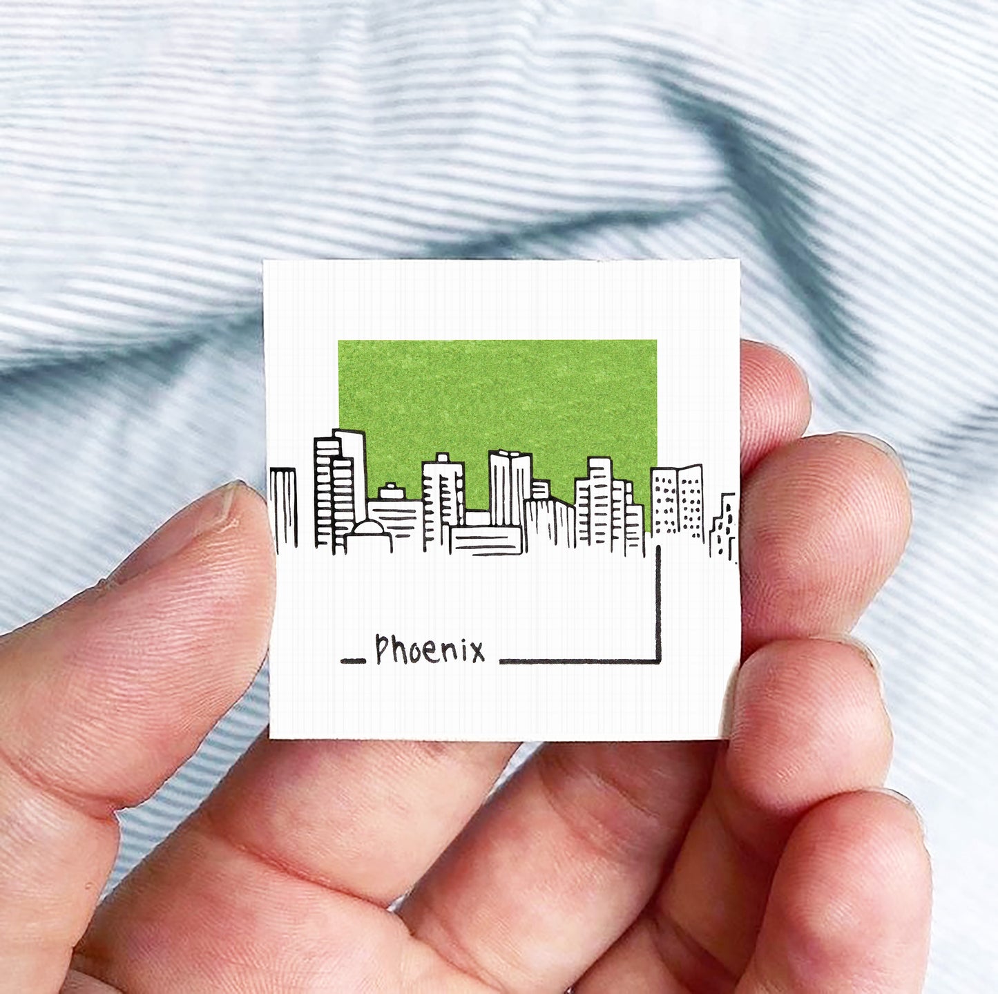 A hand holding a small square with a green city skyline and the word "Phoenix" written below it.