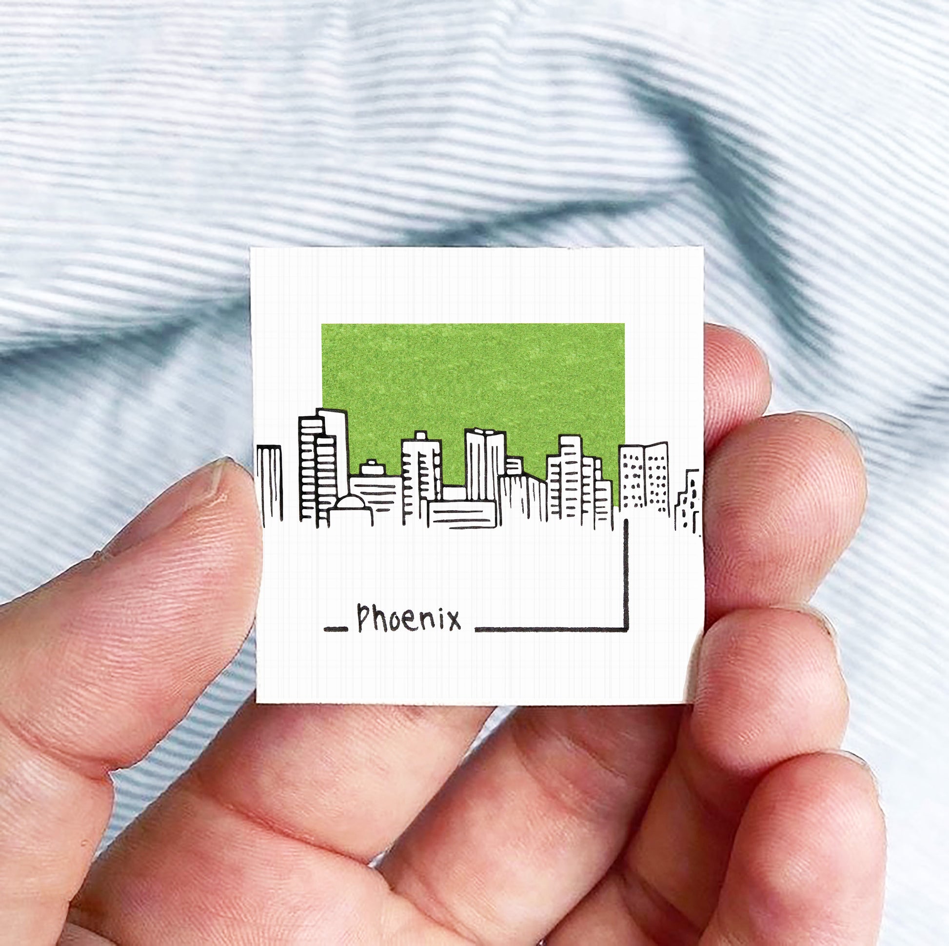 A hand holding a small square with a green city skyline and the word "Phoenix" written below it.