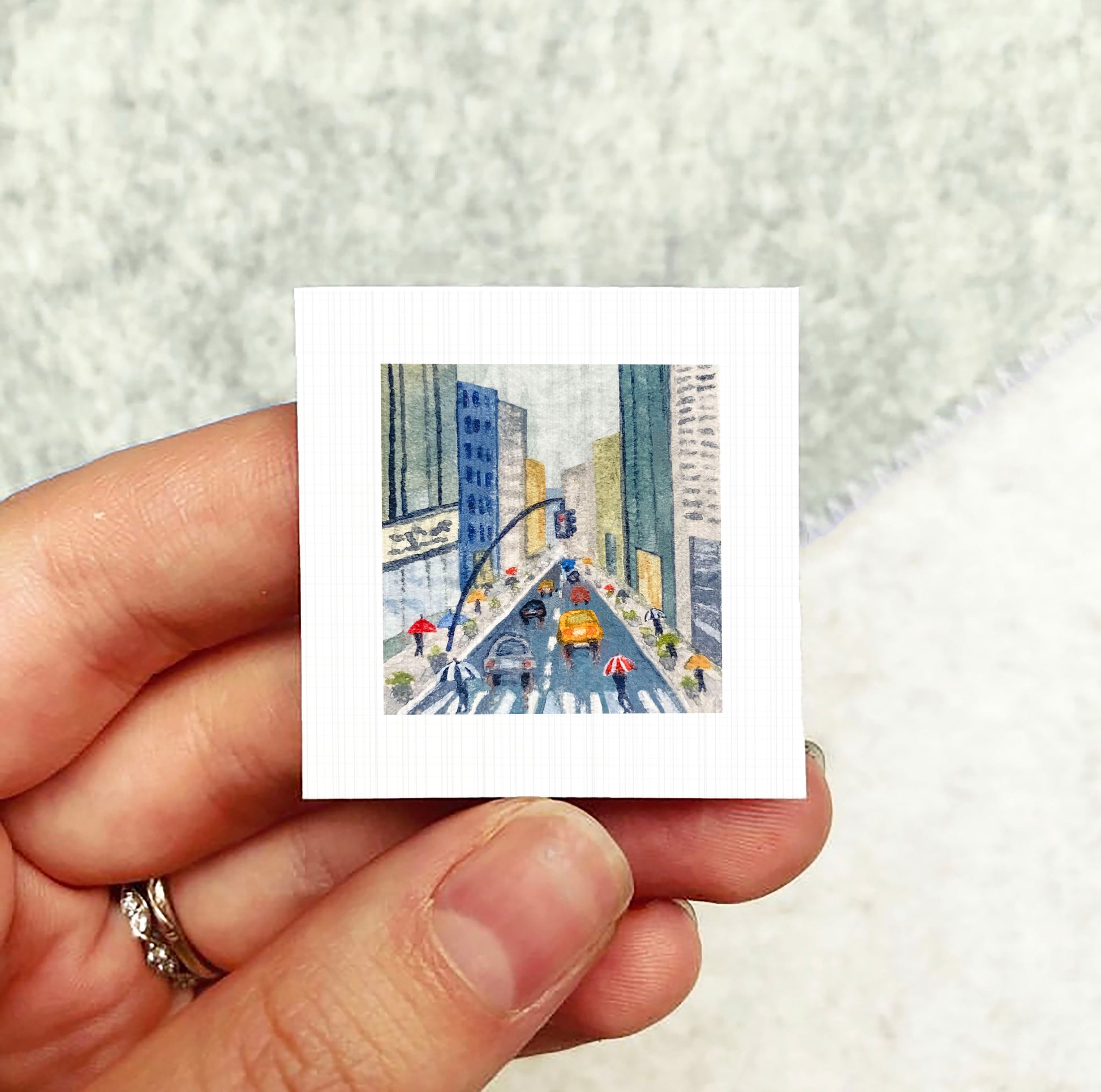 A hand holding a small square artwork depicting a city street scene with cars, pedestrians, and umbrellas.