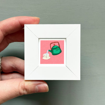 A hand is holding a small white square frame containing an illustration of a green teapot pouring tea into a cup.