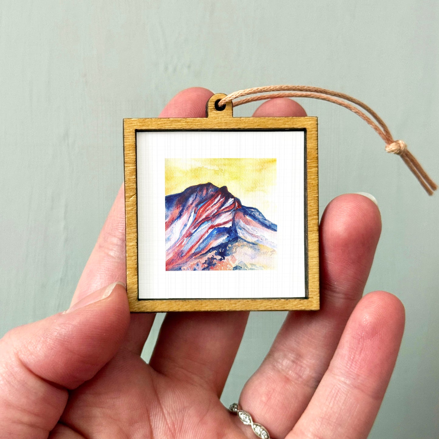 A hand holding a small wooden frame containing a watercolor painting of a mountain landscape.