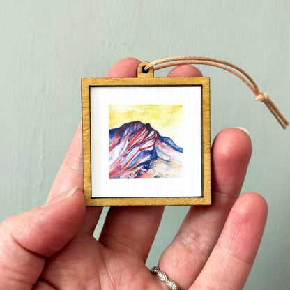 A hand holding a small wooden frame containing a watercolor painting of a mountain landscape.