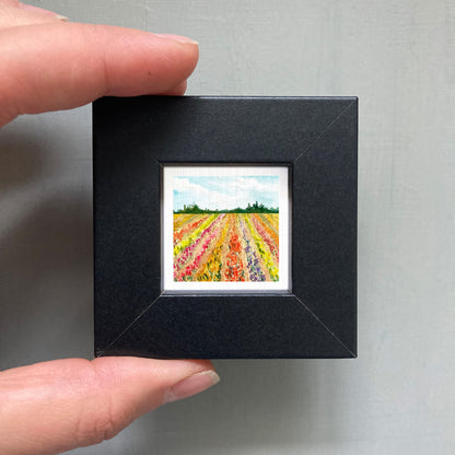 A person's hand holding a small, square-shaped black frame containing a miniature painting of a field of colorful flowers.