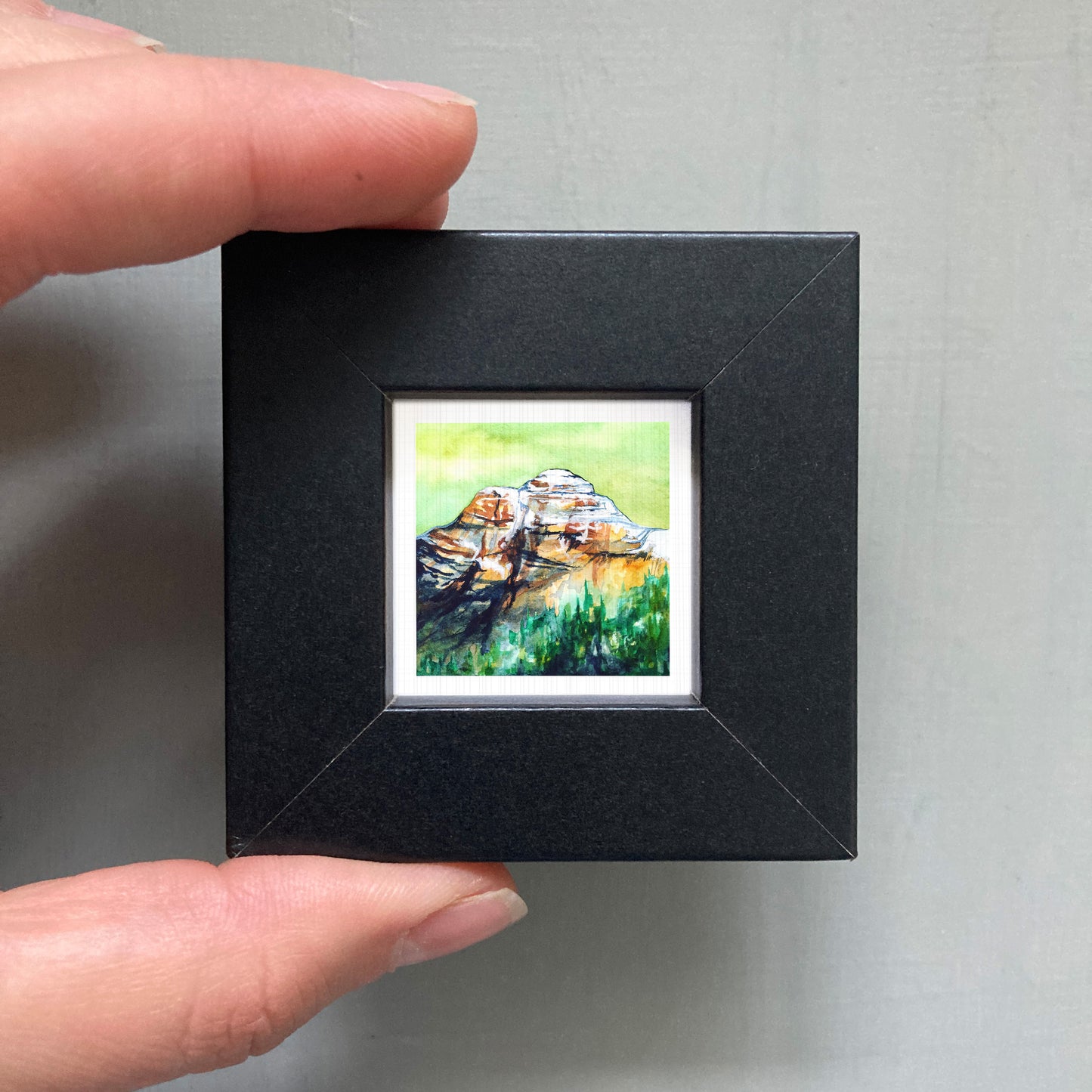 A person's hand holding a small black frame containing a framed watercolor painting of a mountain landscape.