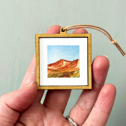 A hand holding a small wooden frame containing a framed watercolor painting of a mountain landscape.