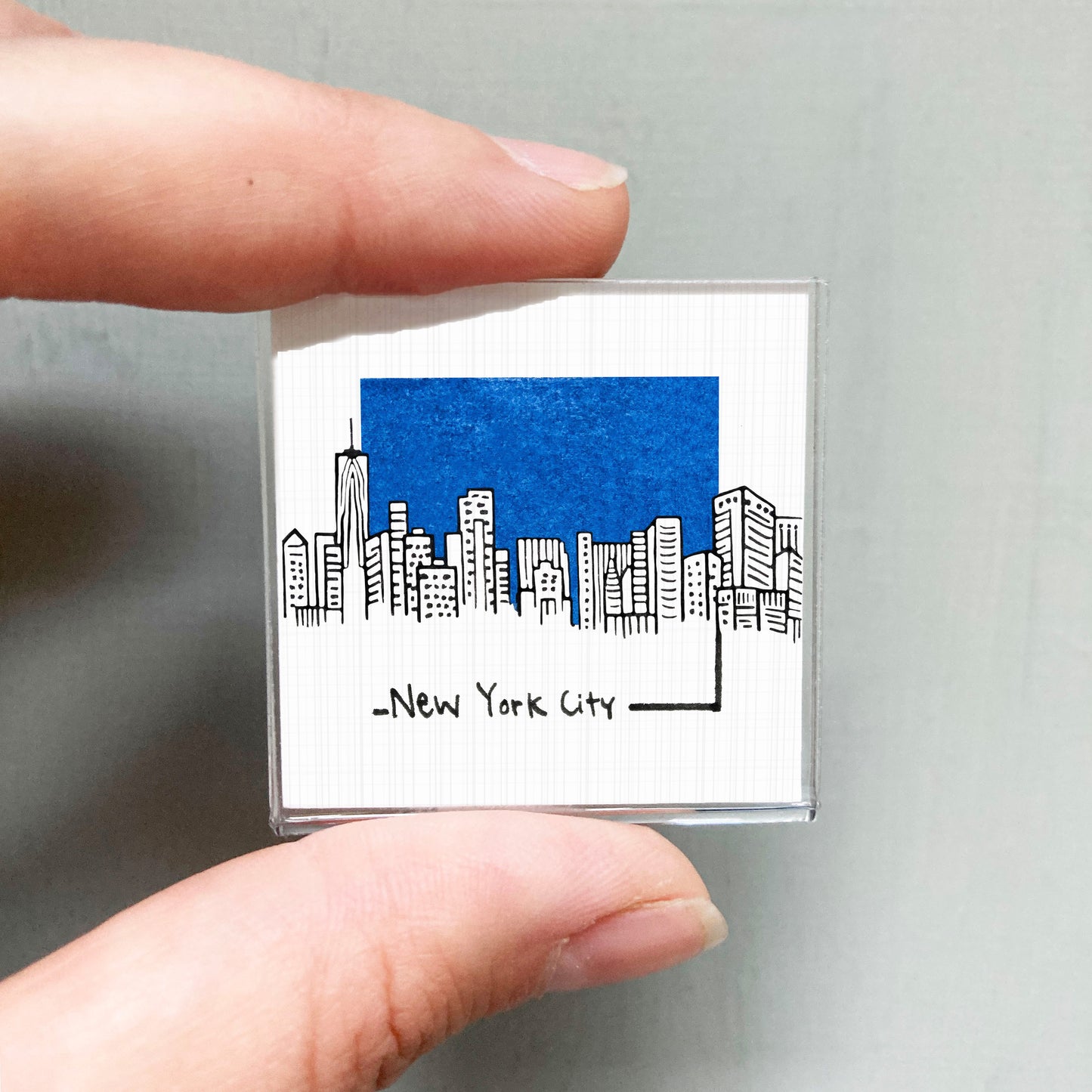 A person's hand holding a small square magnet with a drawing of a city skyline and the text "New York City" written below it.