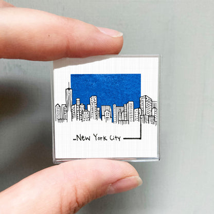 A person's hand holding a small square magnet with a drawing of a city skyline and the text "New York City" written below it.