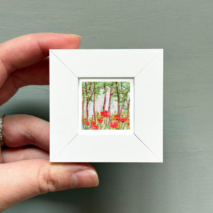 A hand is holding a small white square frame containing a painting of red flowers in a forest.