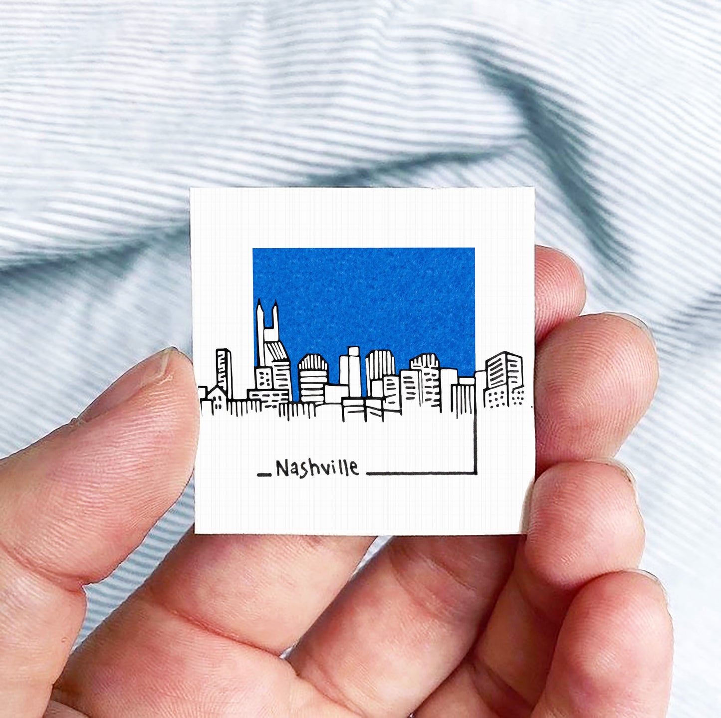 A hand holding a small square-shaped object with a drawing of a city skyline and the word "Nashville" written below it.