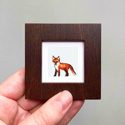 A hand is holding a small wooden frame containing a framed image of a fox.