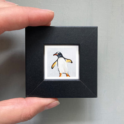 A person's hand is holding a small black frame containing a framed image of a penguin.