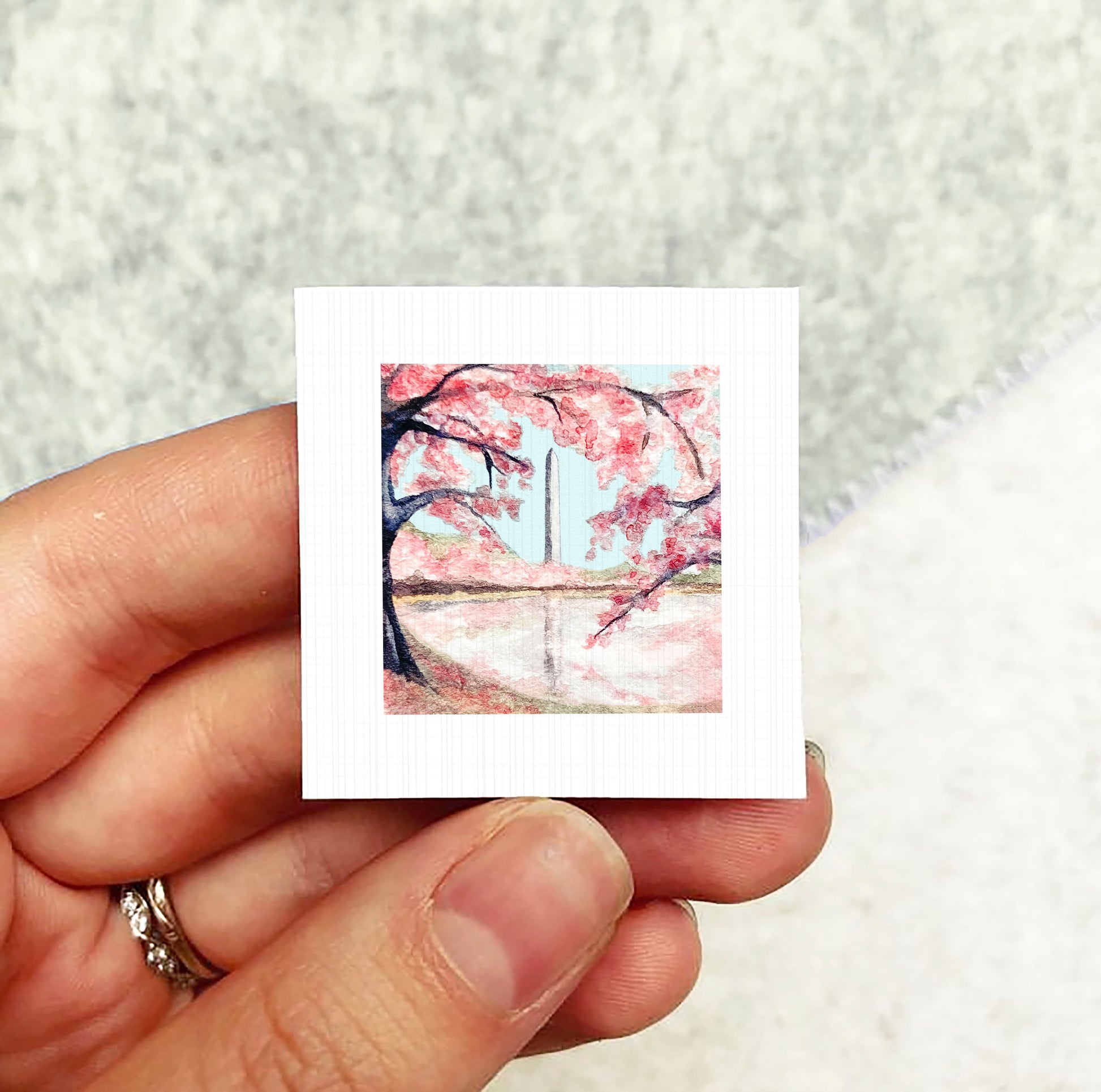 A hand holding a small square picture depicting a cherry blossom tree with pink flowers against a blue sky.