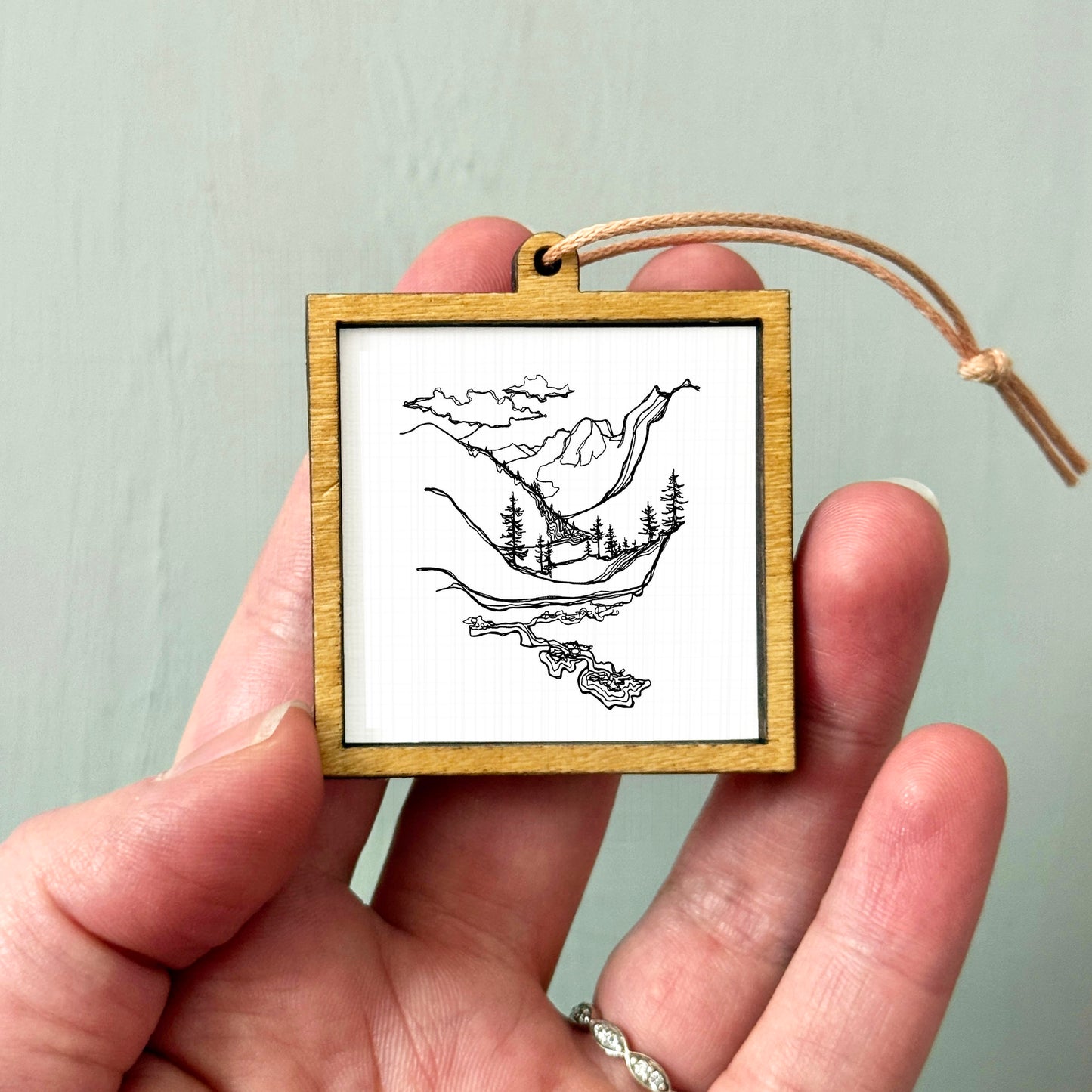 A hand holding a small wooden frame containing a black and white illustration of a landscape with mountains, trees, and a body of water.