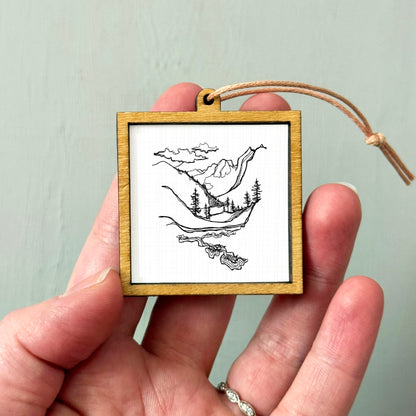 A hand holding a small wooden frame containing a black and white illustration of a landscape with mountains, trees, and a body of water.
