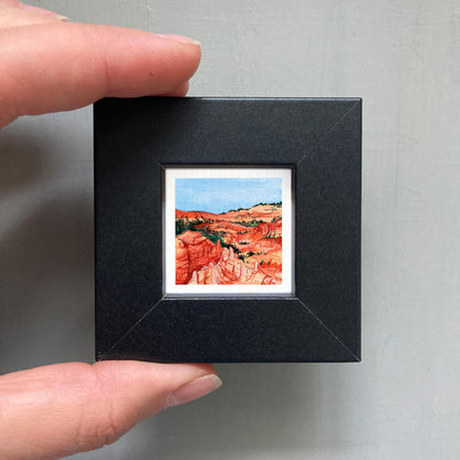 A person's hand holding a small black frame containing a framed watercolor painting of a desert landscape.