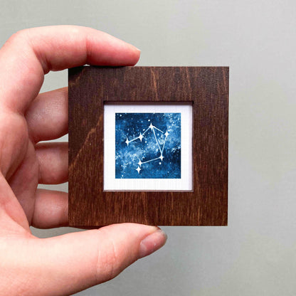 A hand is holding a small wooden frame containing a framed image of a starry night sky.