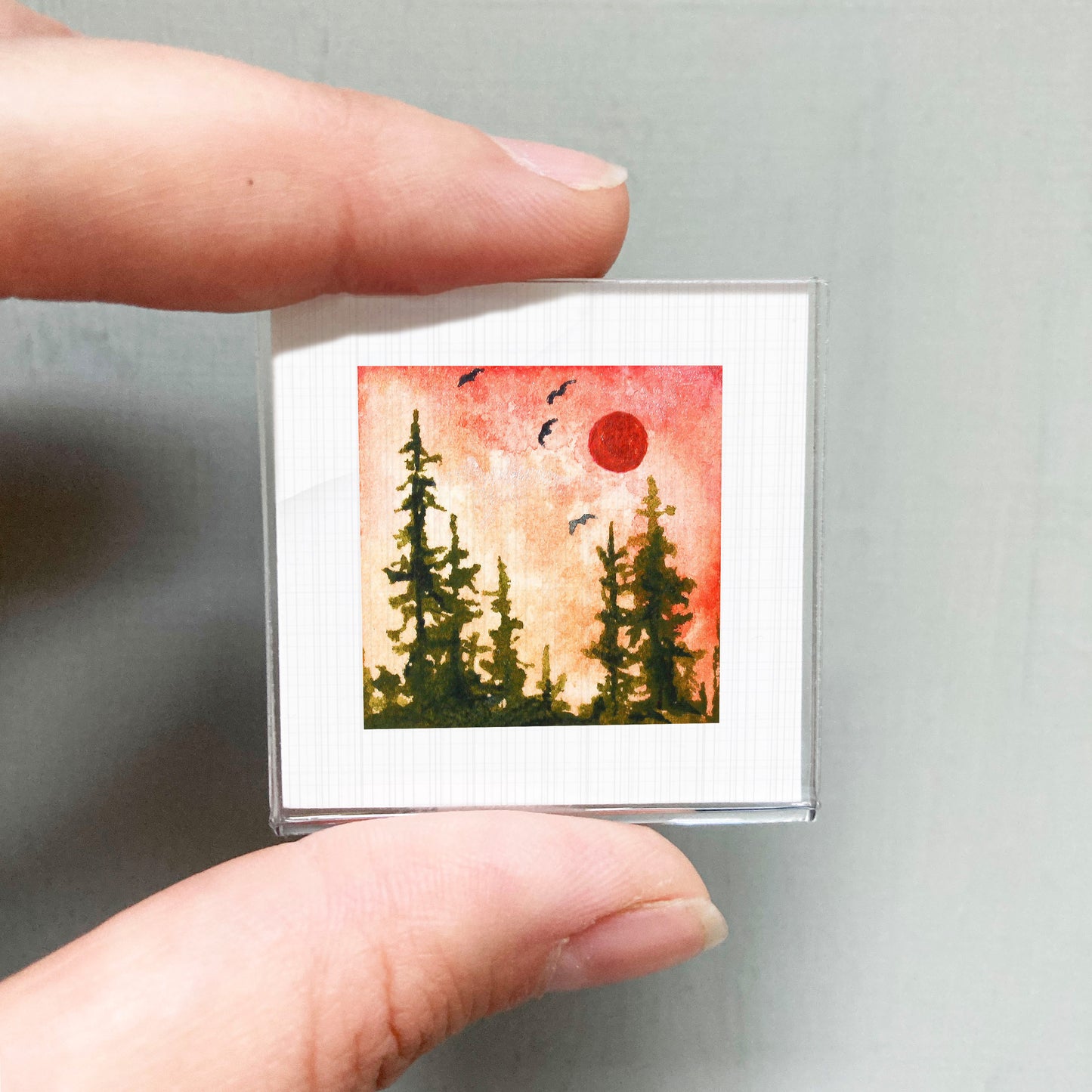 A person's hand is holding a small square picture depicting a forest scene with a red sun in the sky.