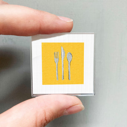 A person's hand holding a small square with a yellow background and three silver utensils, including a fork, knife, and spoon, against a gray background.