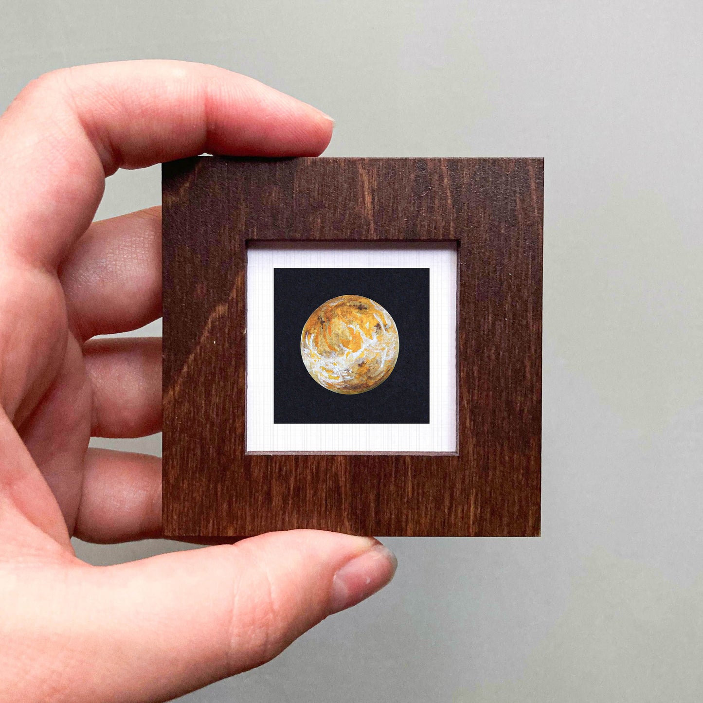 A hand is holding a small wooden frame containing a miniature painting of a planet.