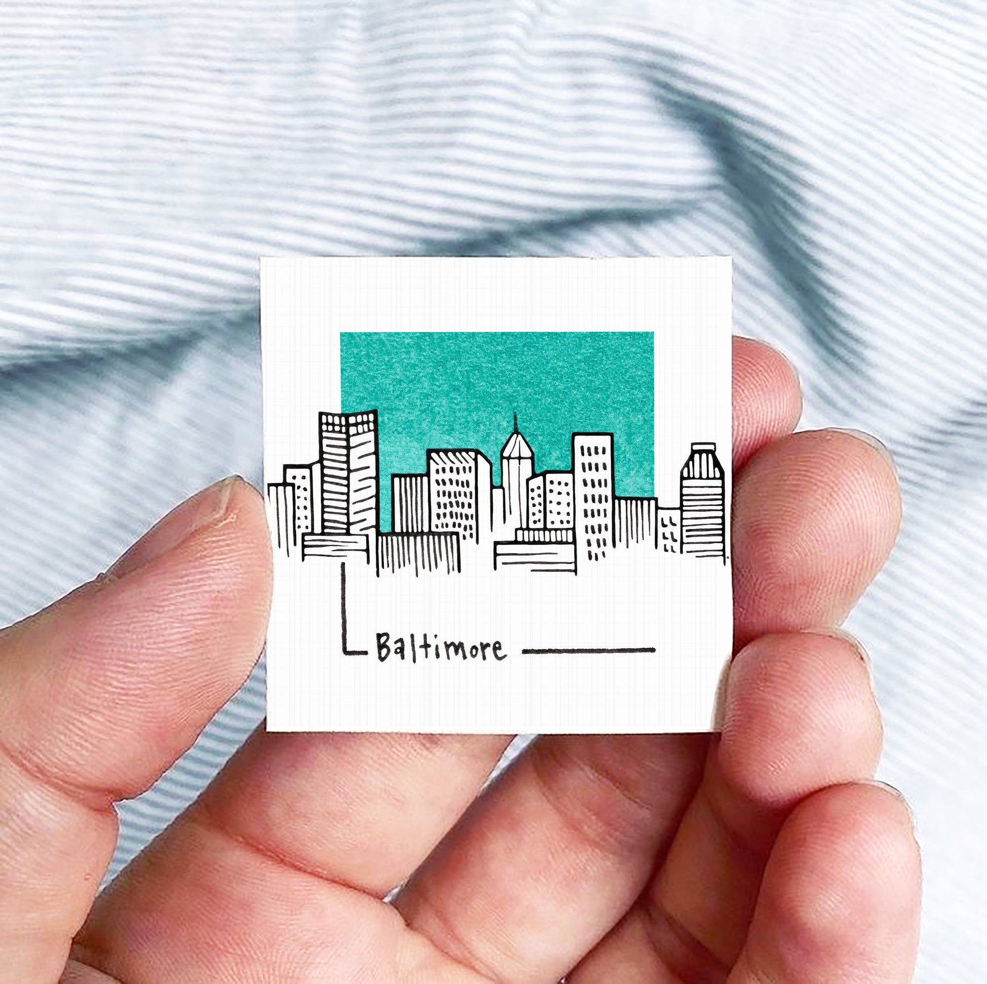 A hand holding a small square-shaped object with a drawing of a city skyline and the word "Baltimore" written on it.