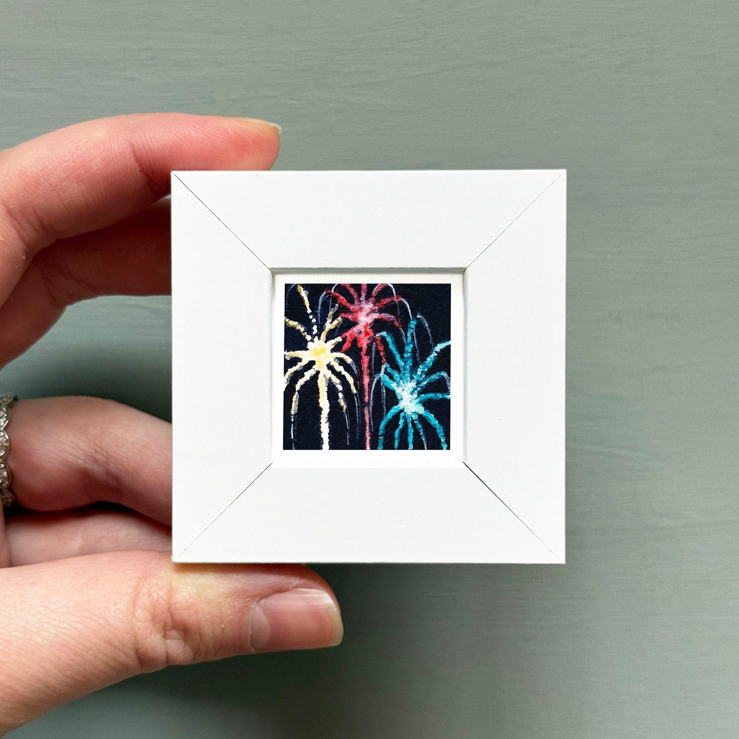 A hand is holding a small white picture frame containing a colorful image of fireworks.
