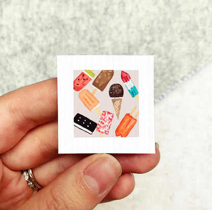 A hand holding a small square card with various colorful ice cream cones and popsicles on it.