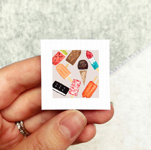 A hand holding a small square card with various colorful ice cream cones and popsicles on it.
