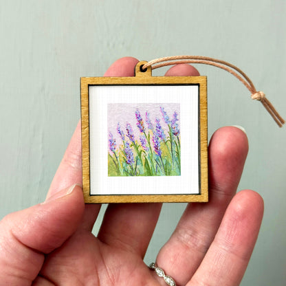 A hand holding a small wooden frame containing a painting of purple flowers.