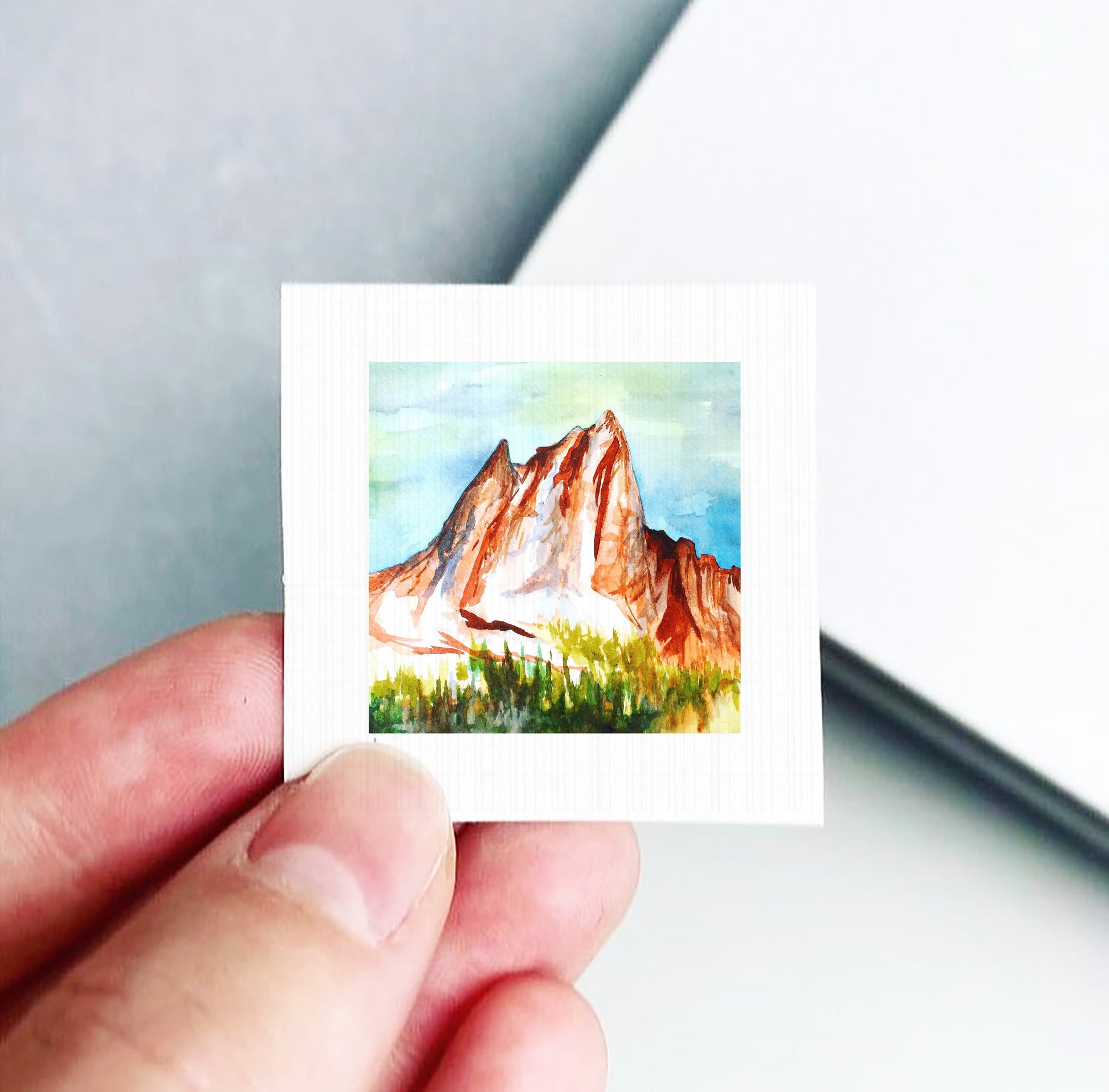 A hand is holding a small square-shaped piece of paper with a painting of a mountain landscape on it.