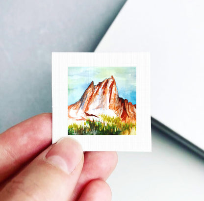 A hand is holding a small square-shaped piece of paper with a painting of a mountain landscape on it.