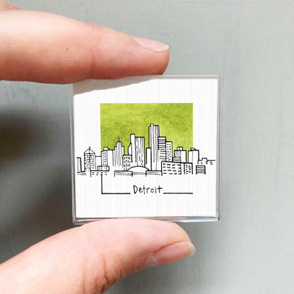 A hand holding a small square magnet with a drawing of a city skyline and the word "Detroit" written on it.