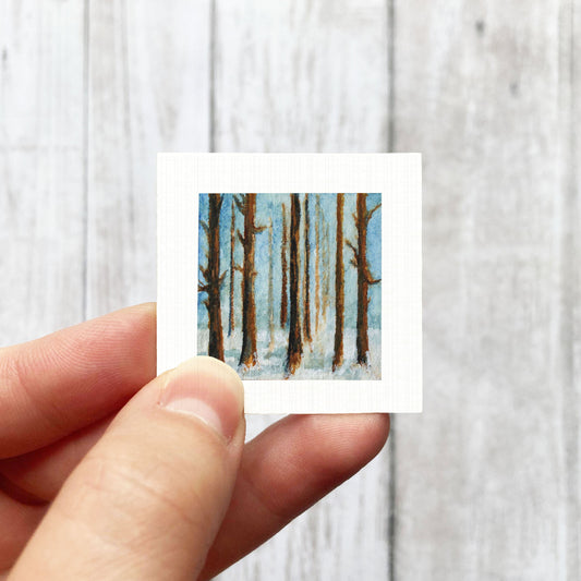 A hand is holding a small square painting of a snowy forest scene.