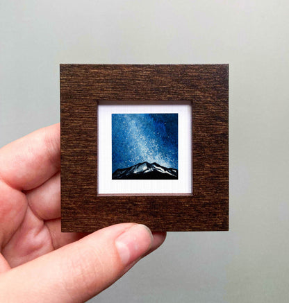 A hand is holding a small wooden frame containing a framed image of a mountain landscape under a starry night sky.