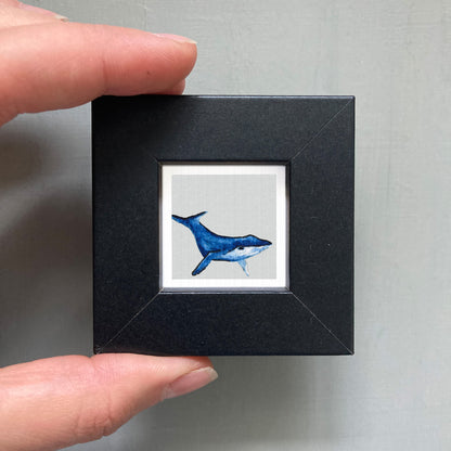 A person's hand is holding a small black frame containing a framed image of a blue whale.
