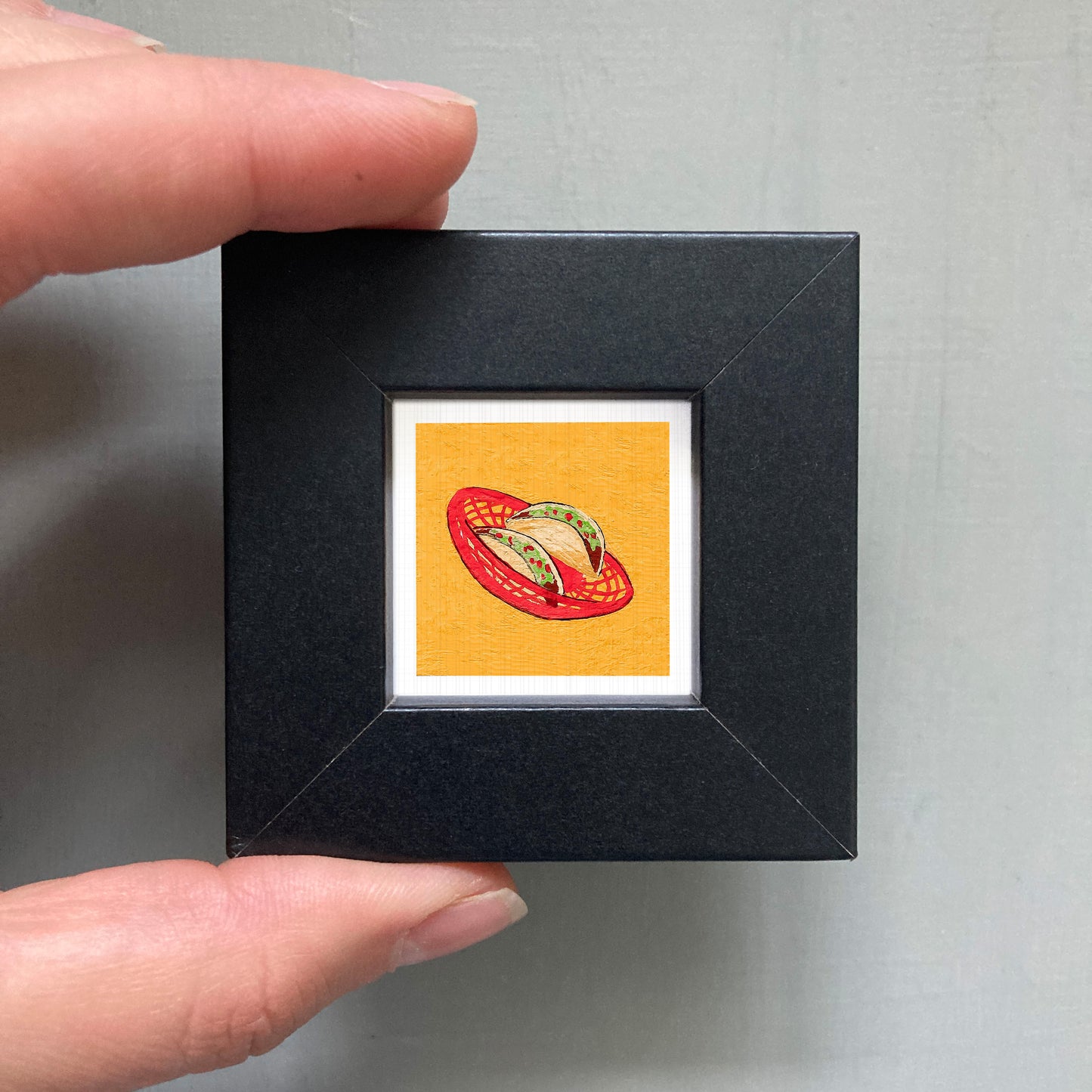 A person's hand is holding a small black frame containing a framed image of a red basket with a yellow interior.