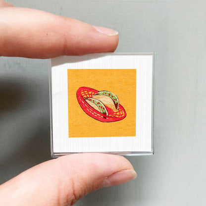 A person's hand holding a small square with a vibrant illustration of a red basket filled with green and yellow objects, resembling a Mexican-style dish.