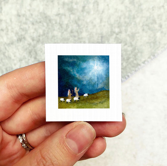 A hand holding a small square picture depicting a pastoral scene with sheep and a starry sky.