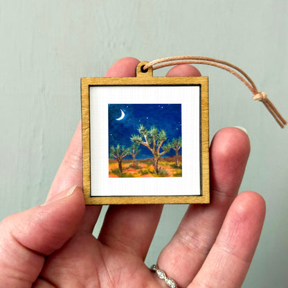 A hand holding a small wooden frame containing a painting of a desert landscape with a crescent moon and stars in the night sky.