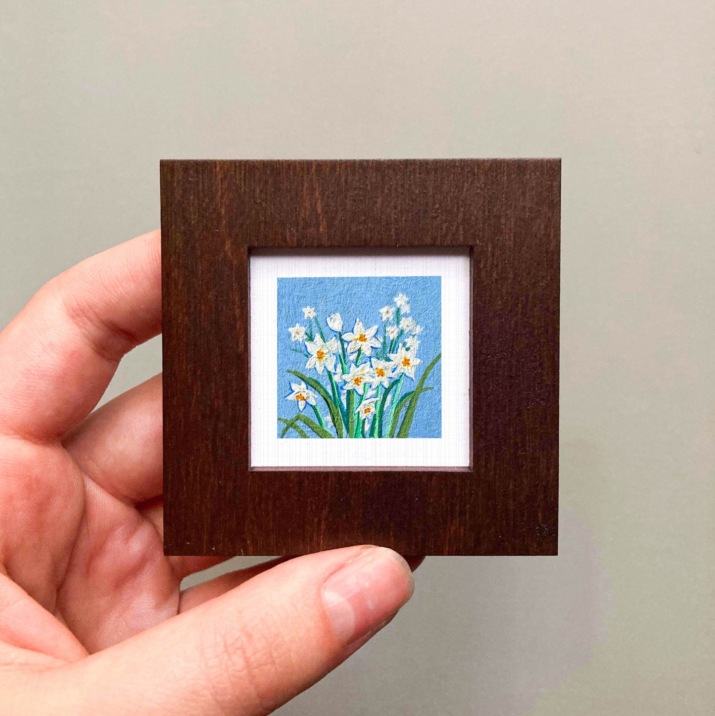 A hand is holding a small wooden frame containing a framed painting of white flowers against a blue background.