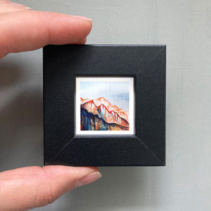A person's hand holding a small black frame containing a framed painting of a mountain landscape.