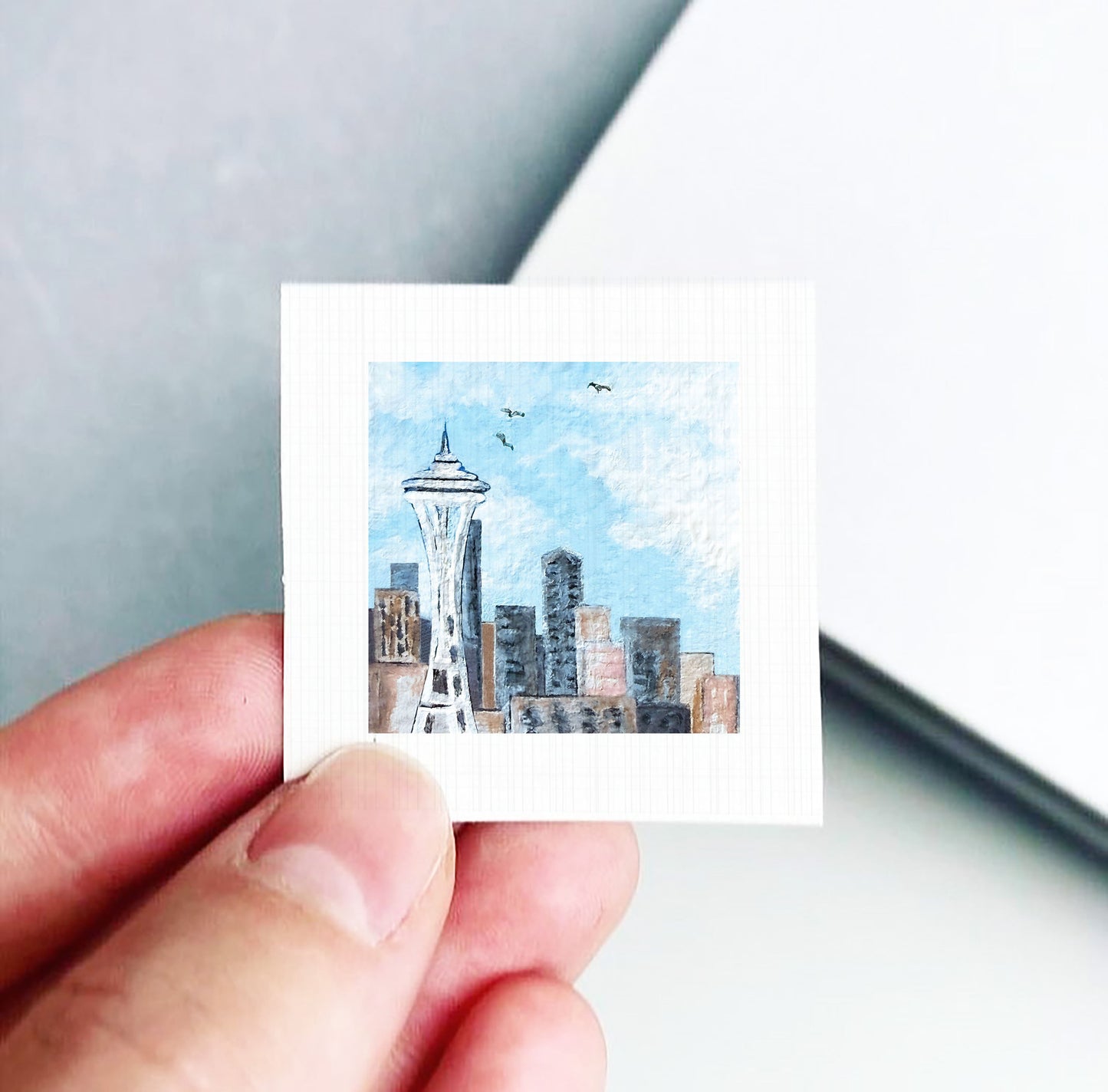 A hand is holding a small square-shaped piece of paper with a painting of the Seattle Space Needle on it.