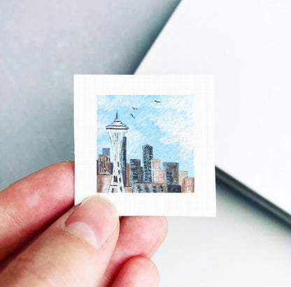 A hand is holding a small square-shaped piece of paper with a painting of the Seattle Space Needle on it.