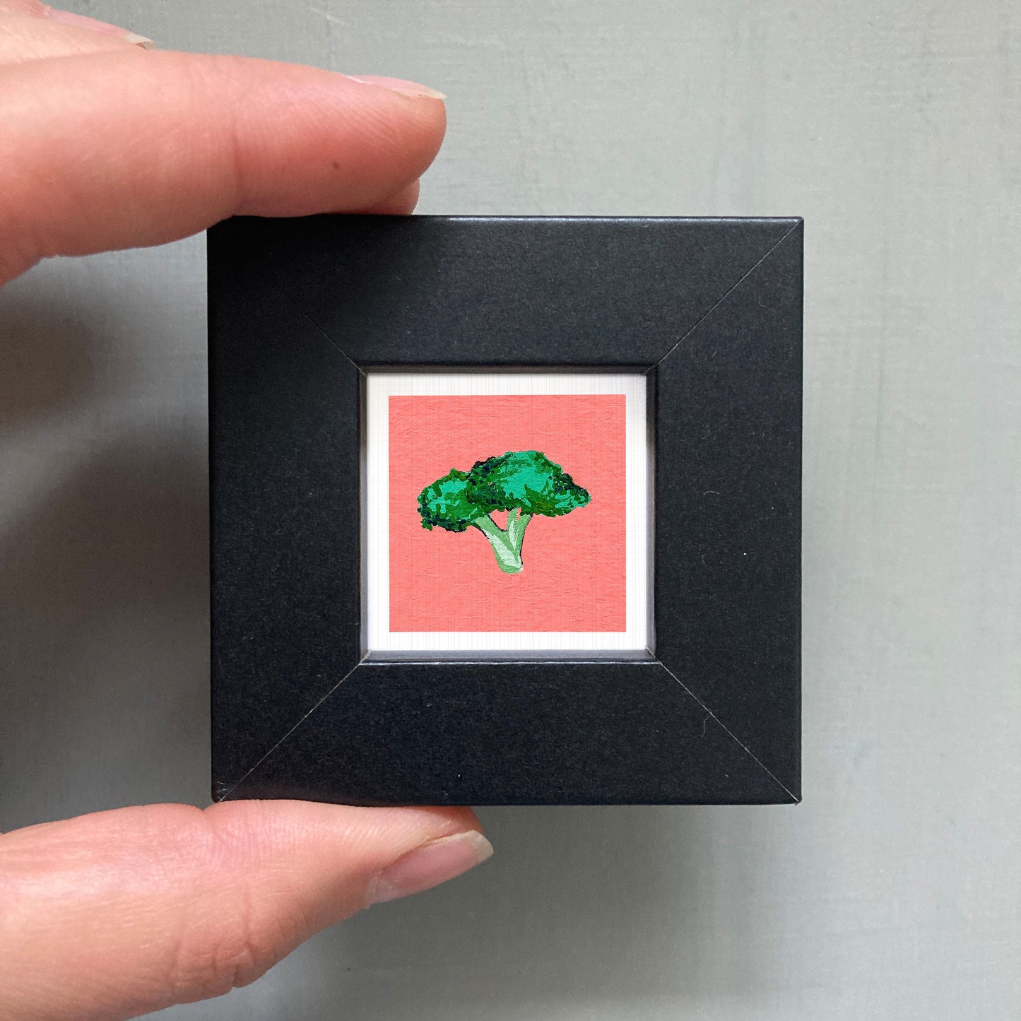 A person's hand is holding a small square picture frame containing a vibrant red background with a green broccoli tree illustration.
