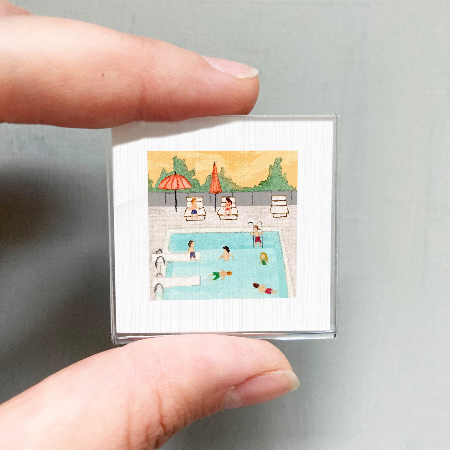 A person's hand is holding a small square picture depicting a swimming pool with people enjoying the water and loungers.