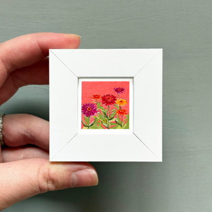 A hand is holding a small white frame containing a vibrant painting of colorful flowers.